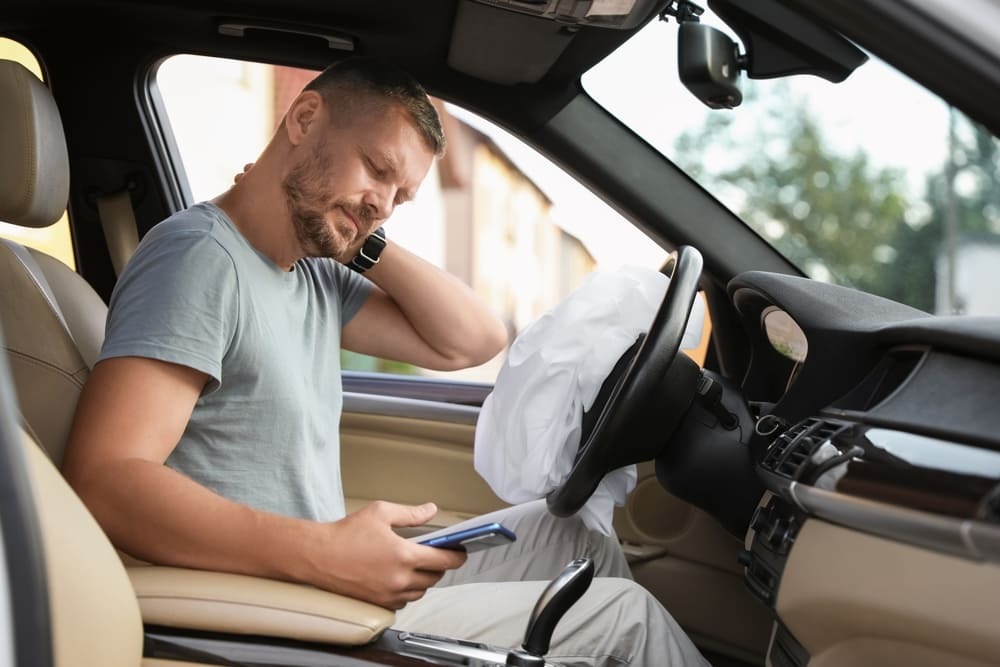 Man in driver's seat after car accident, holding neck in pain while looking at phone with deployed airbag, illustrating common whiplash or neck injury symptoms treated at Multi-Specialty Pain Management in Bronx, NY