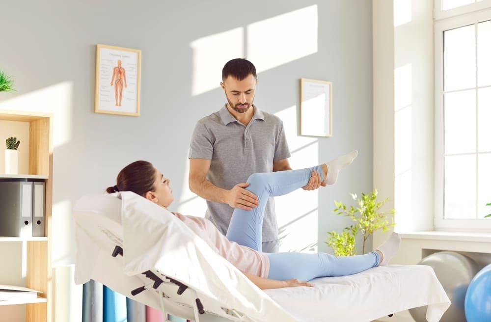 Male physical therapist guiding female patient through passive leg raise and hip flexion exercise on treatment table to improve mobility and reduce pain at Multi-Specialty Pain Management in Bronx, NY
